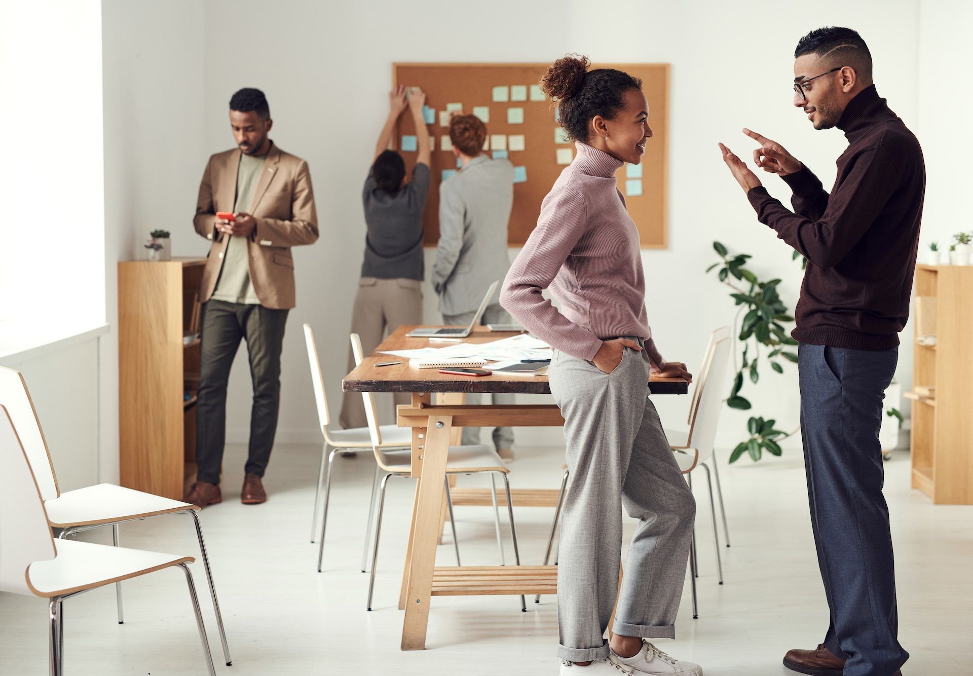 A medium dark skinned person listening to another medium dark skinned person talk while a medium skinned dark person looks at their phone in the background and two other people (both Caucasian) consult a work board .